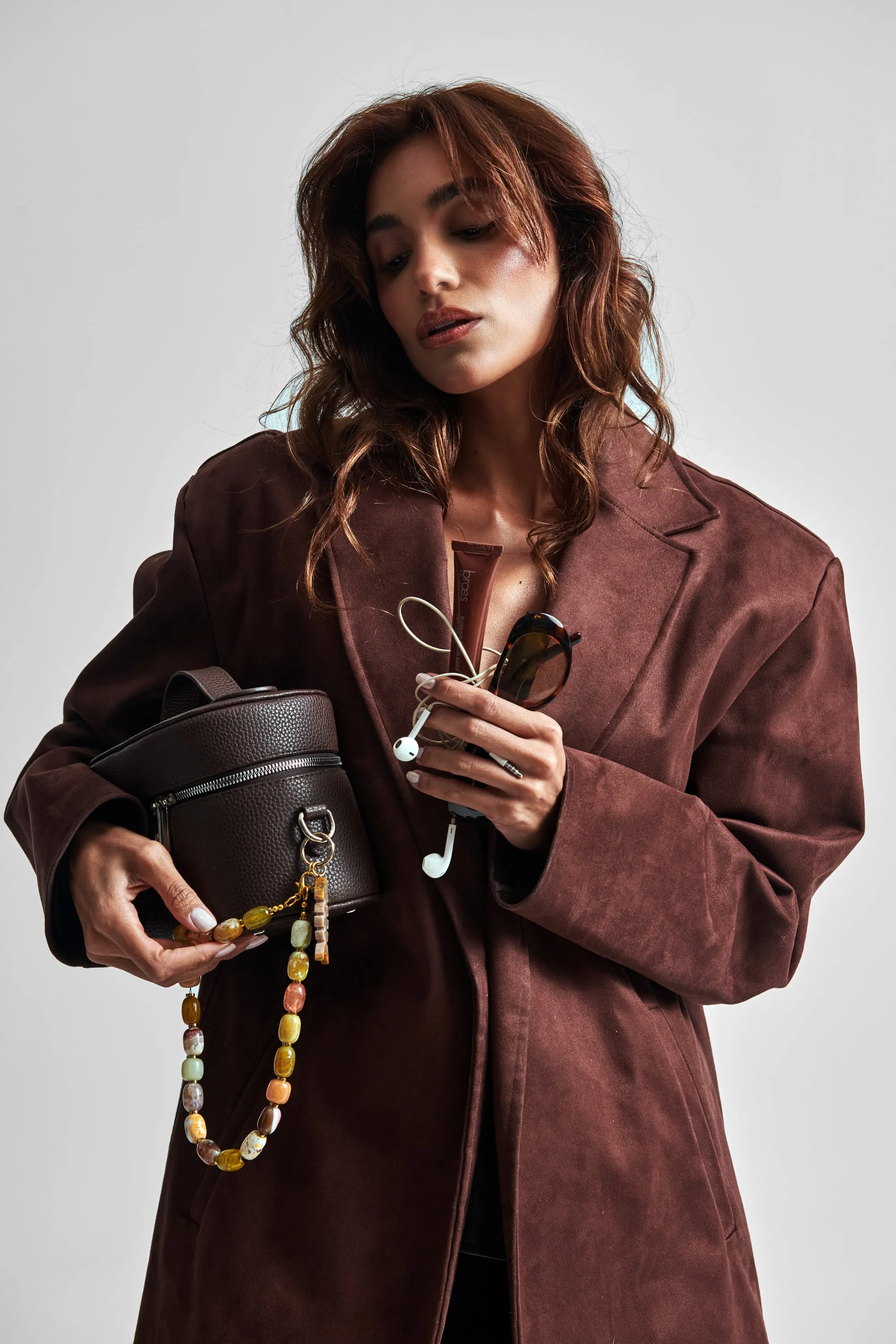 A woman in a brown suede blazer holds a dark brown vanity bag with a beaded strap, plus sunglasses and earphones.