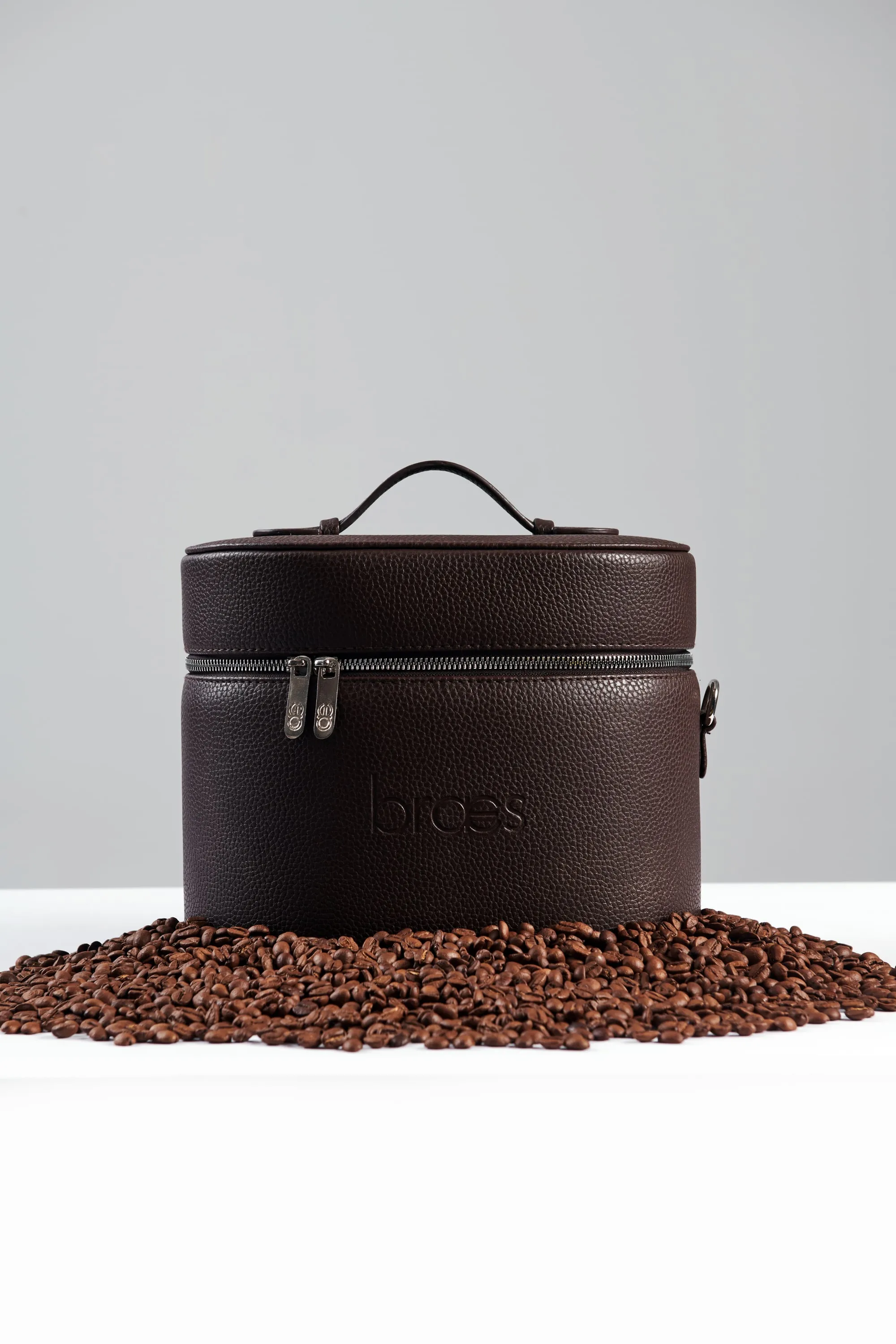 A dark brown leather vanity bag embossed with the braes logo, set on a bed of coffee beans.