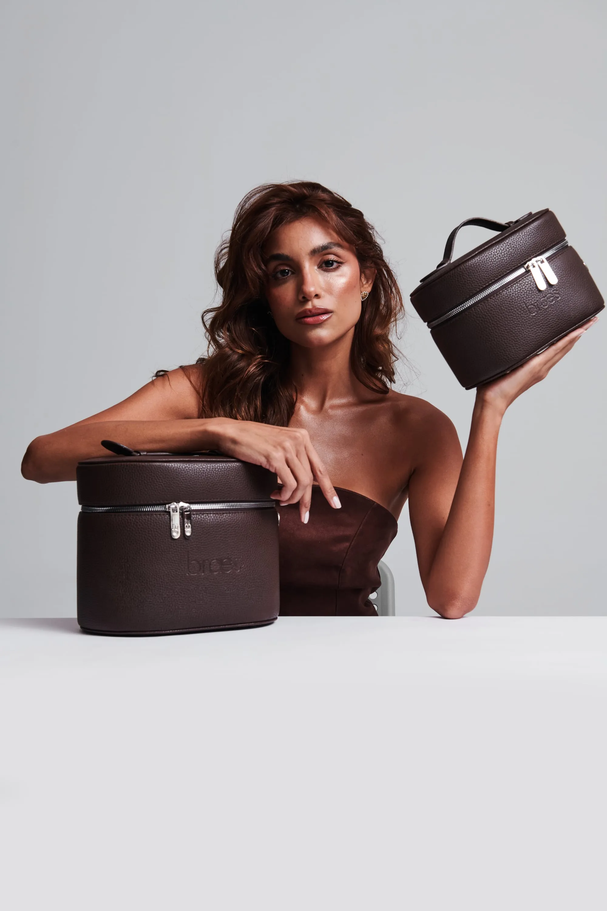 A woman in a strapless brown top leans on one dark brown vanity bag at a table and holds up a second.