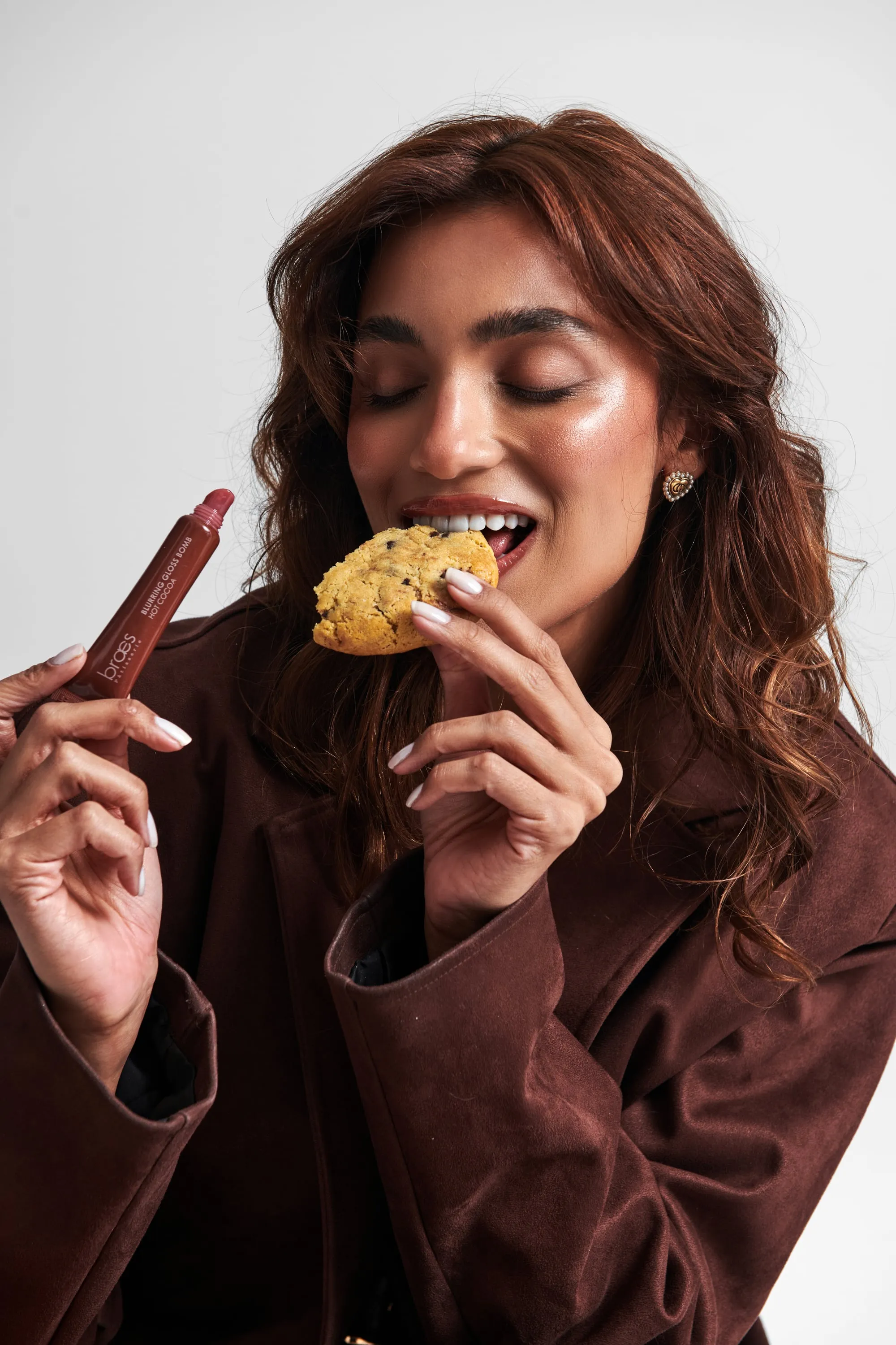 A woman with closed eyes bites a cookie while holding up a brown Blurring Gloss Bomb lip gloss tube.