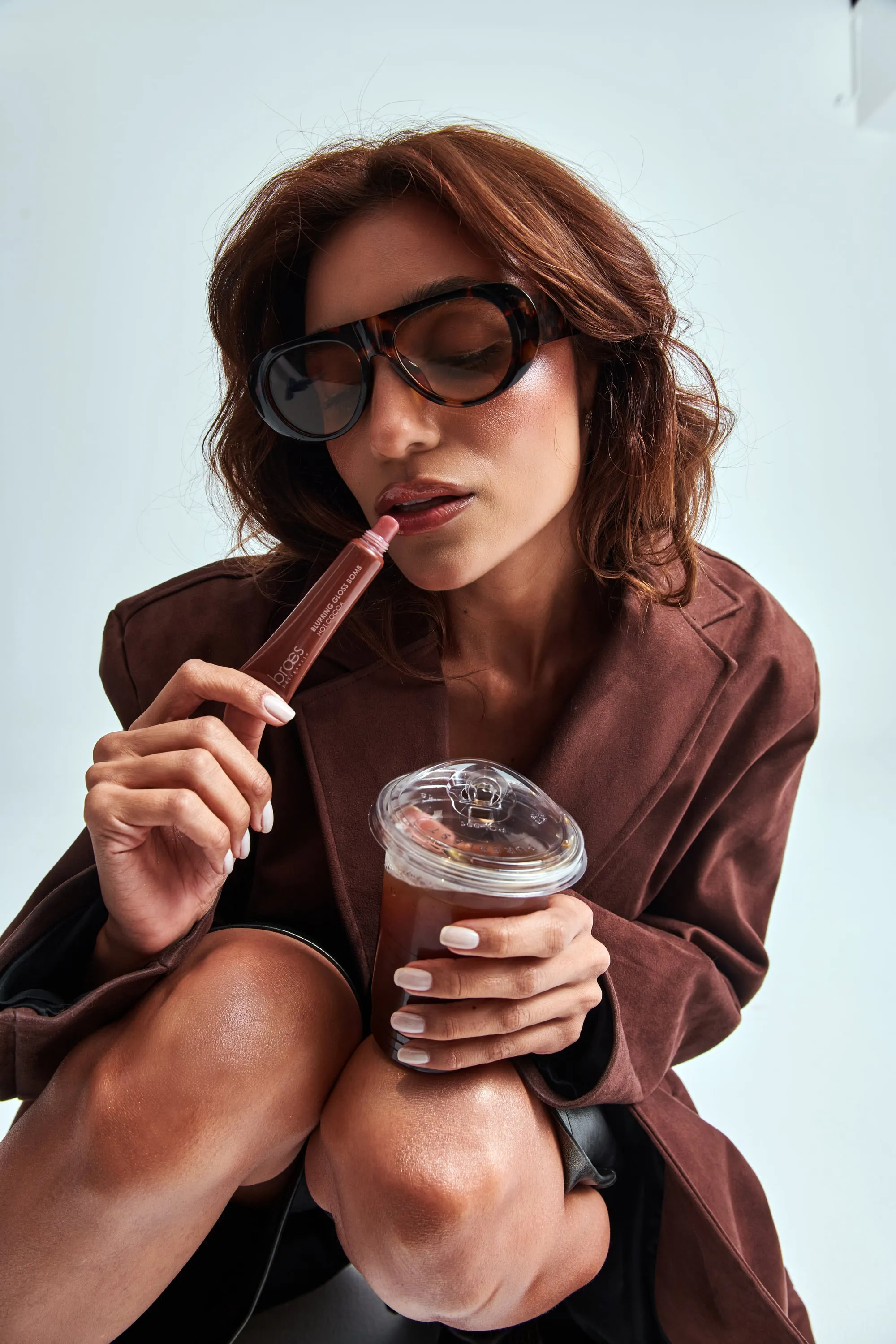 A woman in tortoiseshell sunglasses applies a brown lip gloss while holding an iced drink.