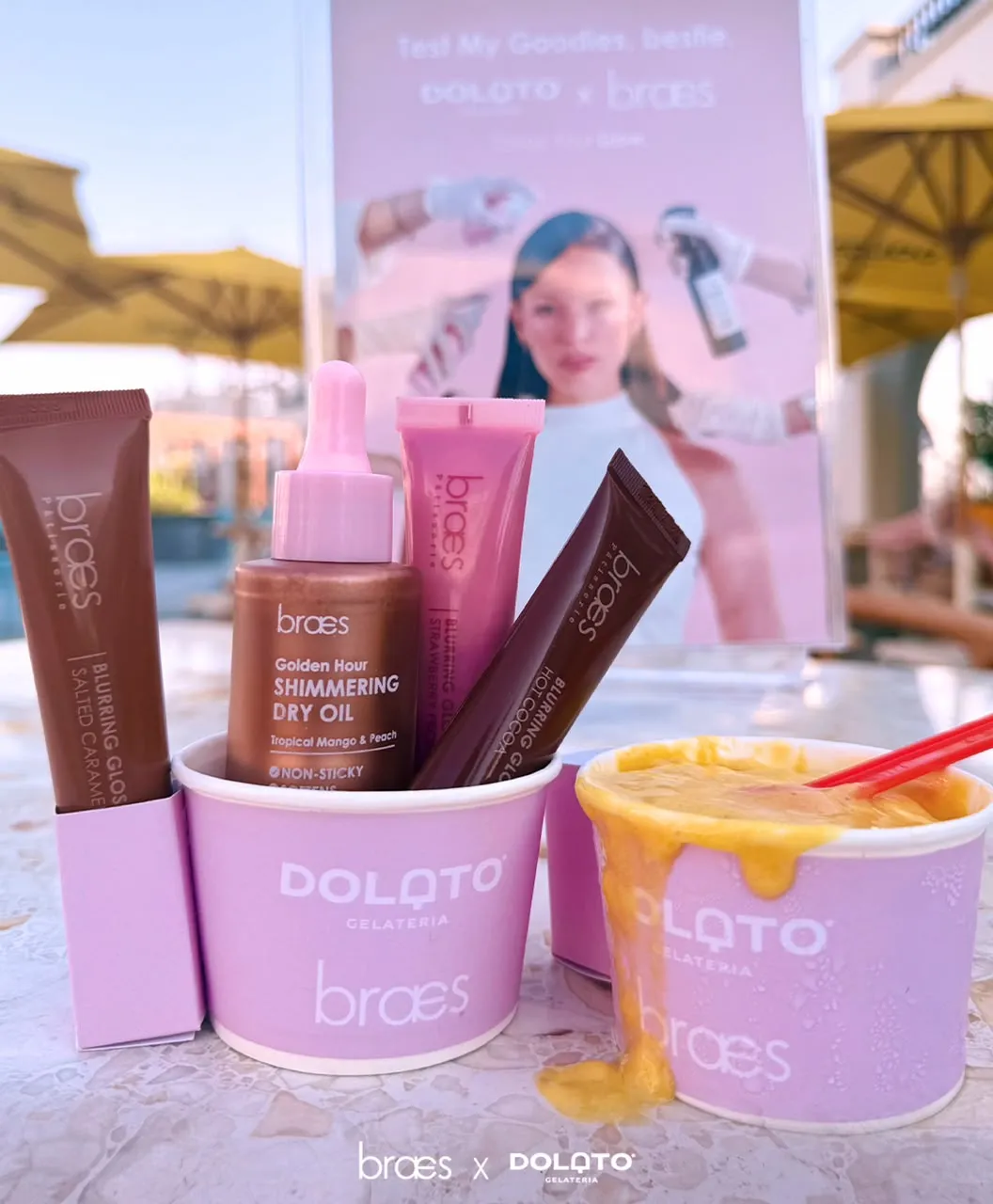 Braes lip glosses and dry oil in Dolato gelato cups beside a mango gelato, under a sign reading Test My Goodies, bestie.
