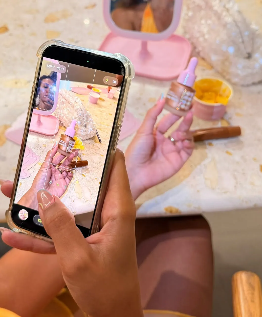 A hand films a small rose-gold serum bottle on a phone at a table with a pink mirror and gelato.